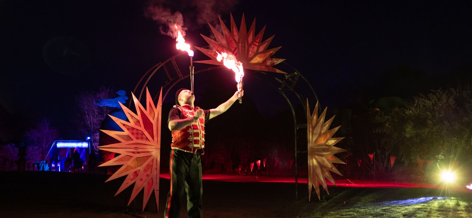 man fire breathing at night time with colourful lights in the background.